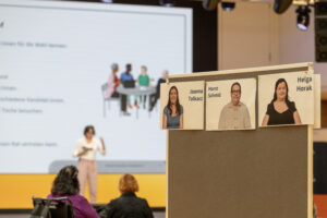 Fotos von Kandidat:innen an einer Pinnwand. Im Hintergrund sieht man die Bühne.