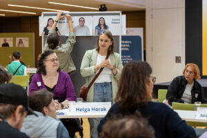 Eine Tisch, eine Frau sitzt, die andere steht. Im Hintergrund pinnt ein Mann etwas an eine Pinnwand.