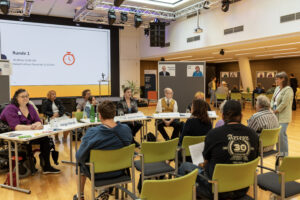 Die kandidat:innen sitzen am Anfang des Raums an einem Tisch. man sieht das Publikum von hinten.