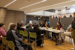 Mehrere Personen sitzen auf Stühlen und blicken Richtung mehreren Personen, die an einem Tisch sitzen.