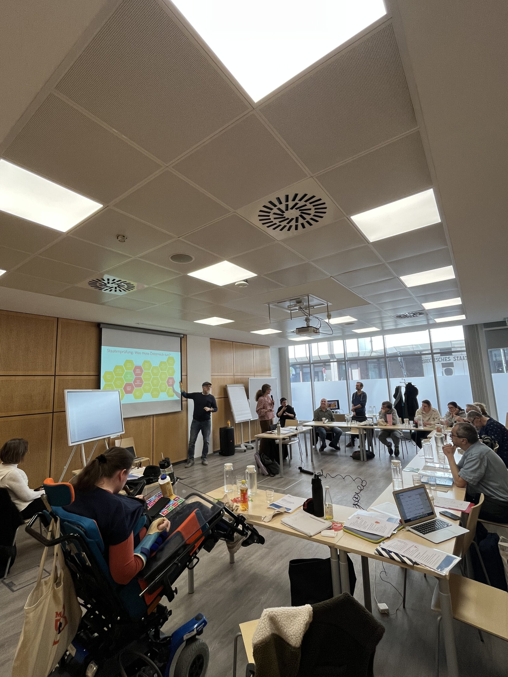 Menschen sitzen in U-Formation an Tischen und hören einem Vortrag zu