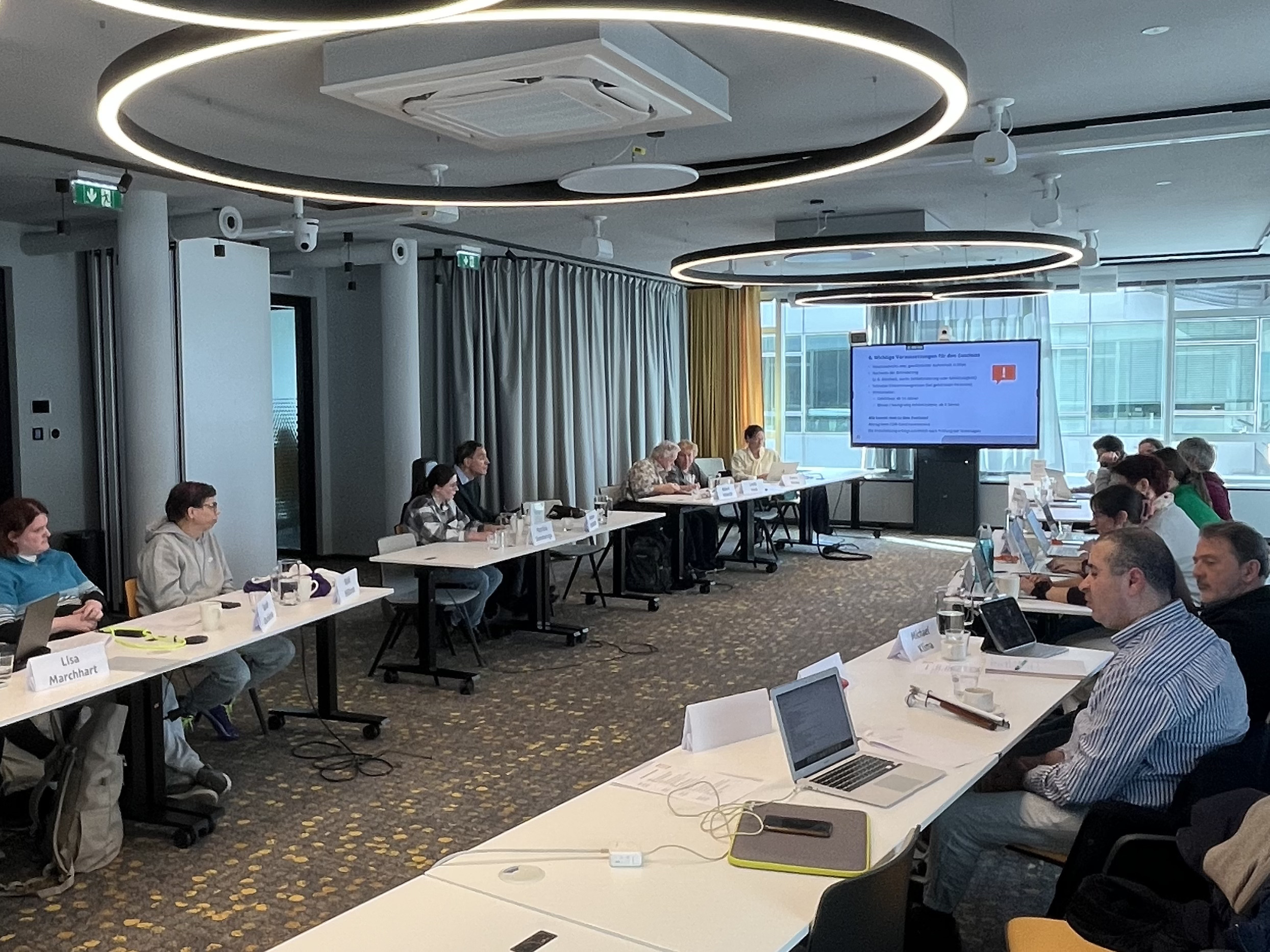 Menschen halten eine Sitzung ab. Sie sitzen an zwei langen Tischen gegenüber.